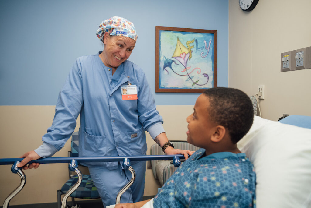 A smiling nurse in blue scrubs and a patterned scrub cap leans over a hospital bed to talk to a young boy.
