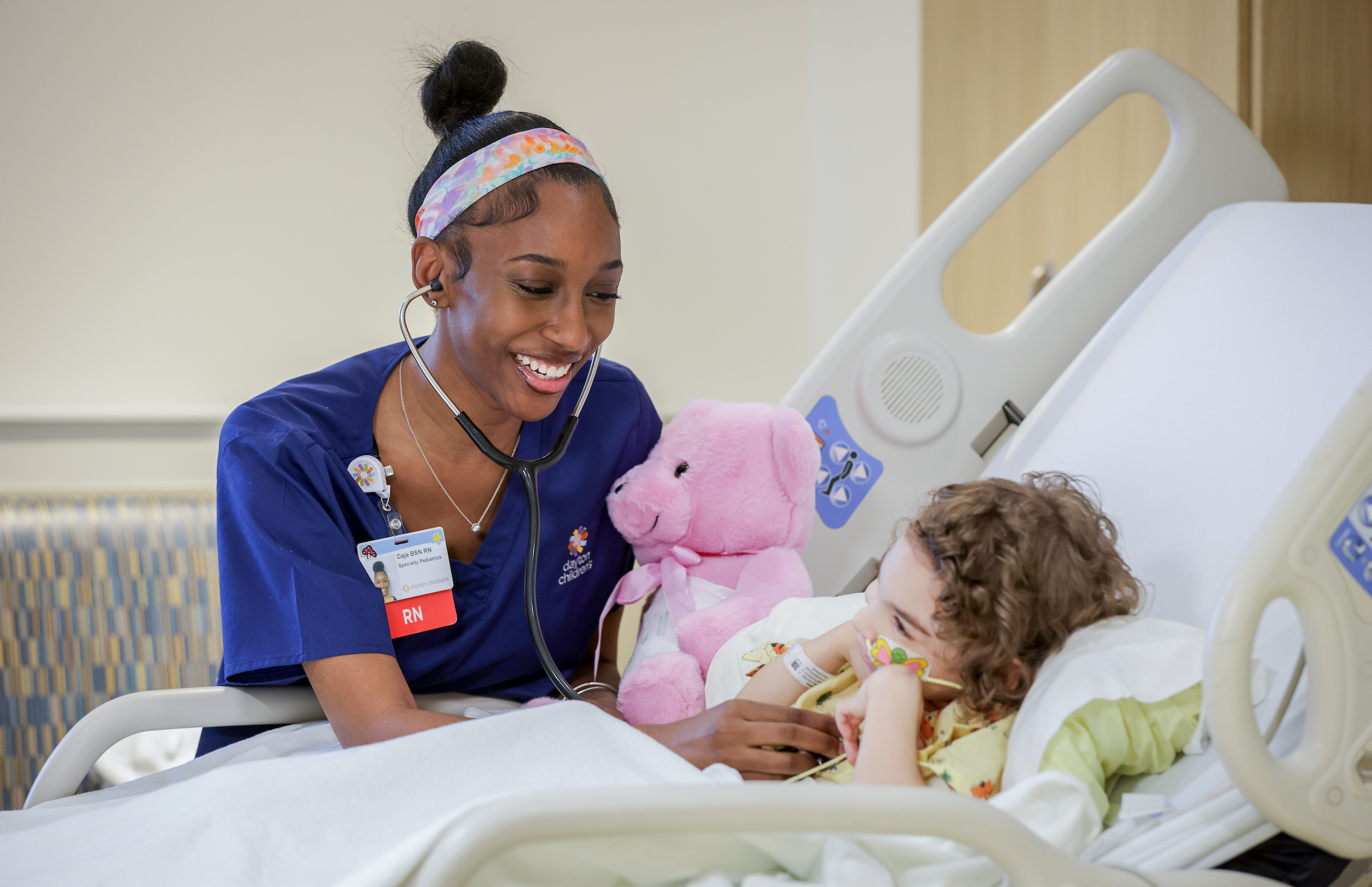 A smiling Black nurse in blue scrubs and a headband interacts with a young patient and a pink teddy bear in a hospital bed.
