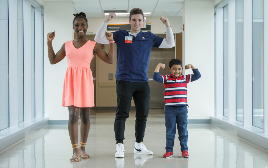 Three people, a smiling teenage girl, a male nurse, and a young boy, flex their biceps in a bright hospital hallway.