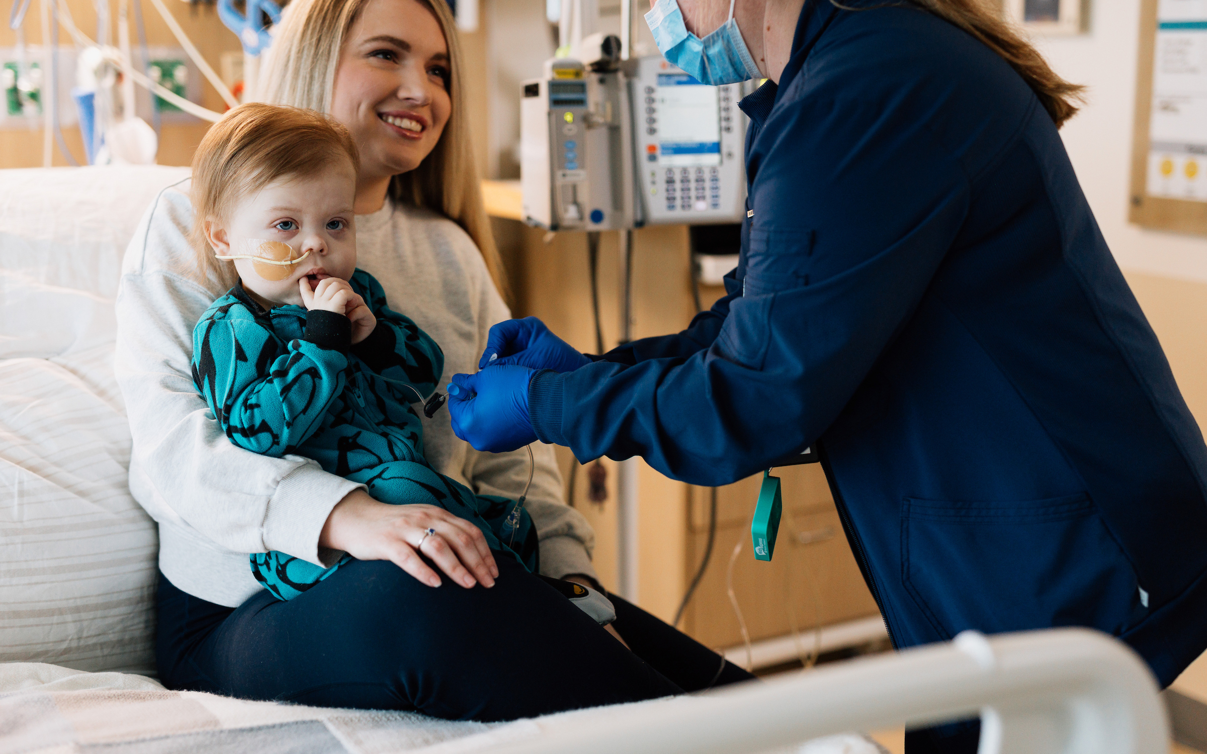 A mother smiles while holding a baby with a feeding tube, as a nurse in blue scrubs and a mask adjusts the baby's tubing.
