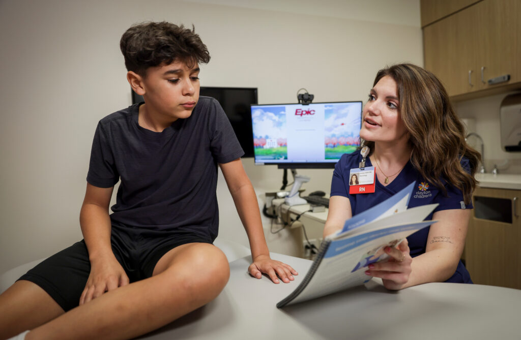 A female nurse or medical professional in blue scrubs speaks to a teenage boy sitting on an exam table while holding a brochure.

