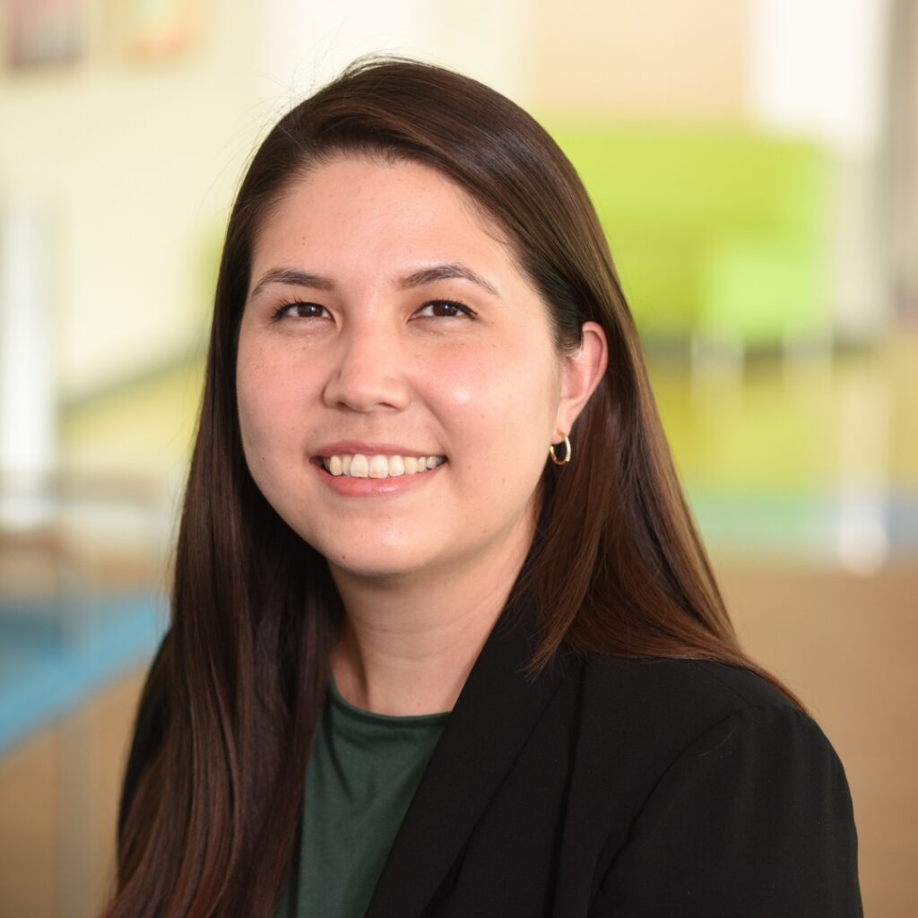 A professional headshot of a smiling woman with long dark hair, wearing a black blazer over a green top.