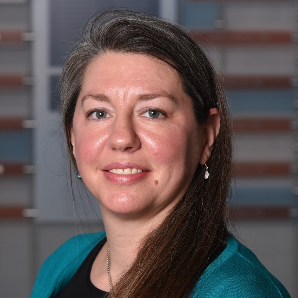 A professional headshot of a smiling woman with long hair wearing a teal top and a necklace.