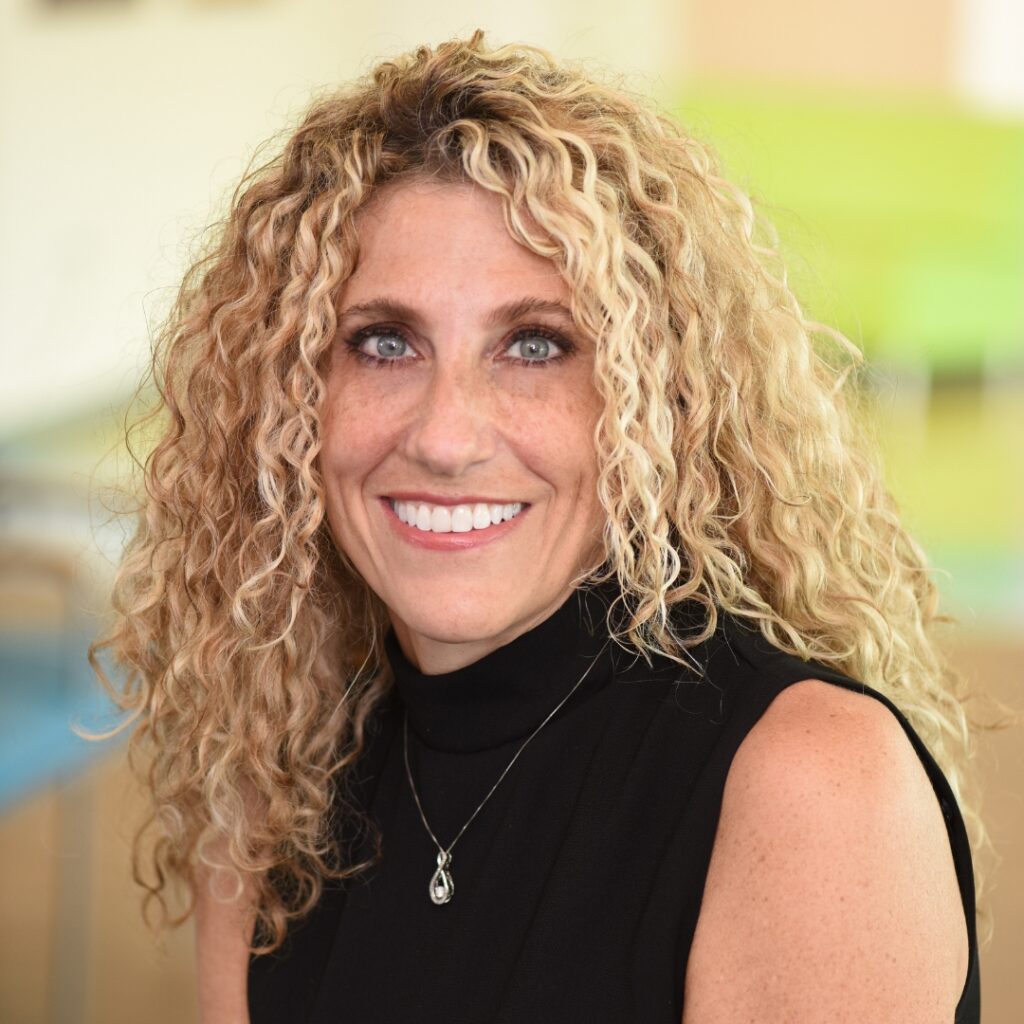 A professional headshot of a smiling woman with blonde hair, wearing a black top and a pendant necklace.
