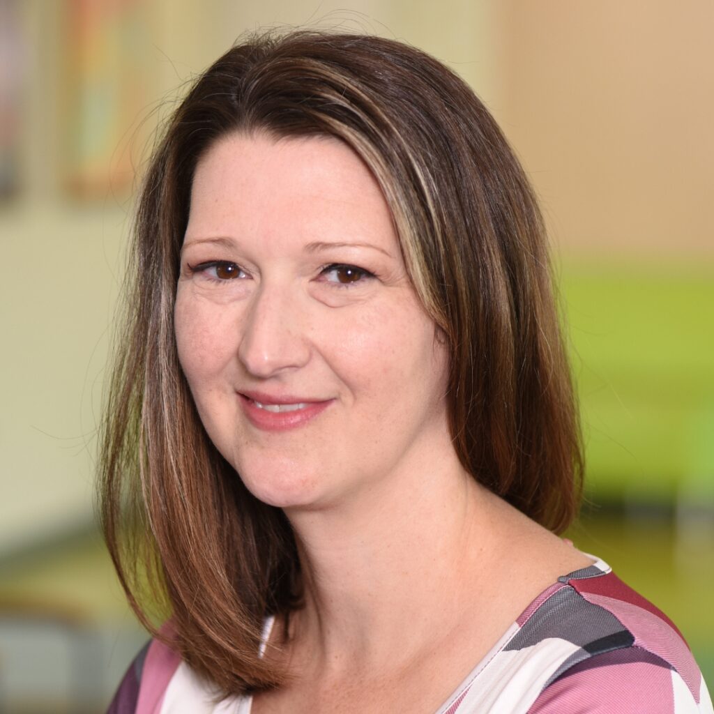 A professional headshot of a smiling woman with shoulder-length brown hair wearing a plaid top