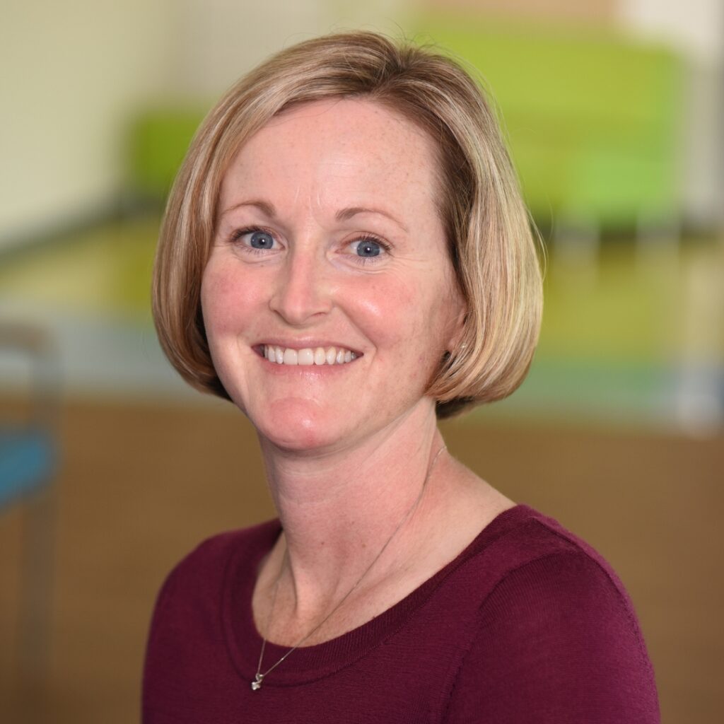 A professional headshot of a smiling woman with short blonde hair, wearing a burgundy top and a necklace.