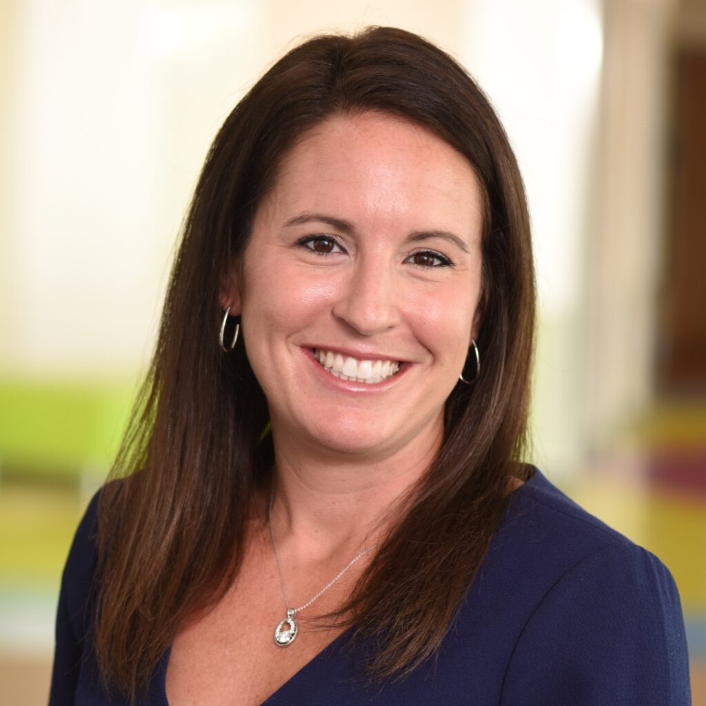 A professional headshot of a smiling woman with long brown hair, wearing a dark blue top and a pendant necklace with hoop earrings.