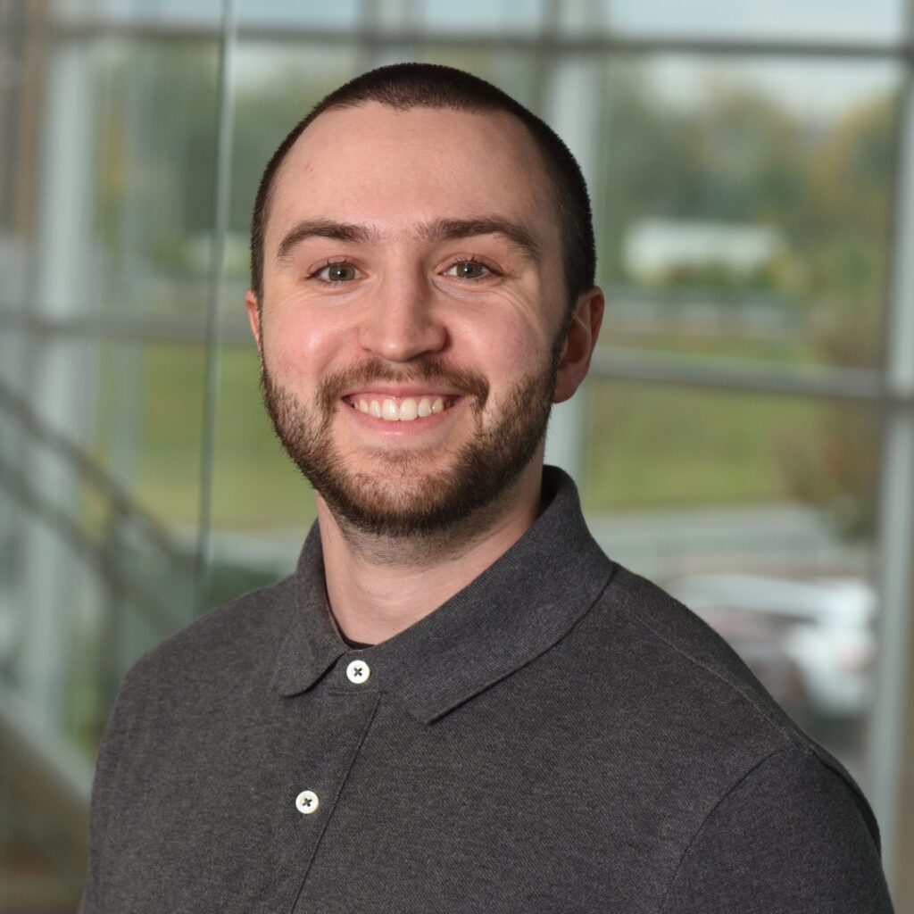 A professional headshot of a smiling man with short brown hair, wearing a grey collared shirt.