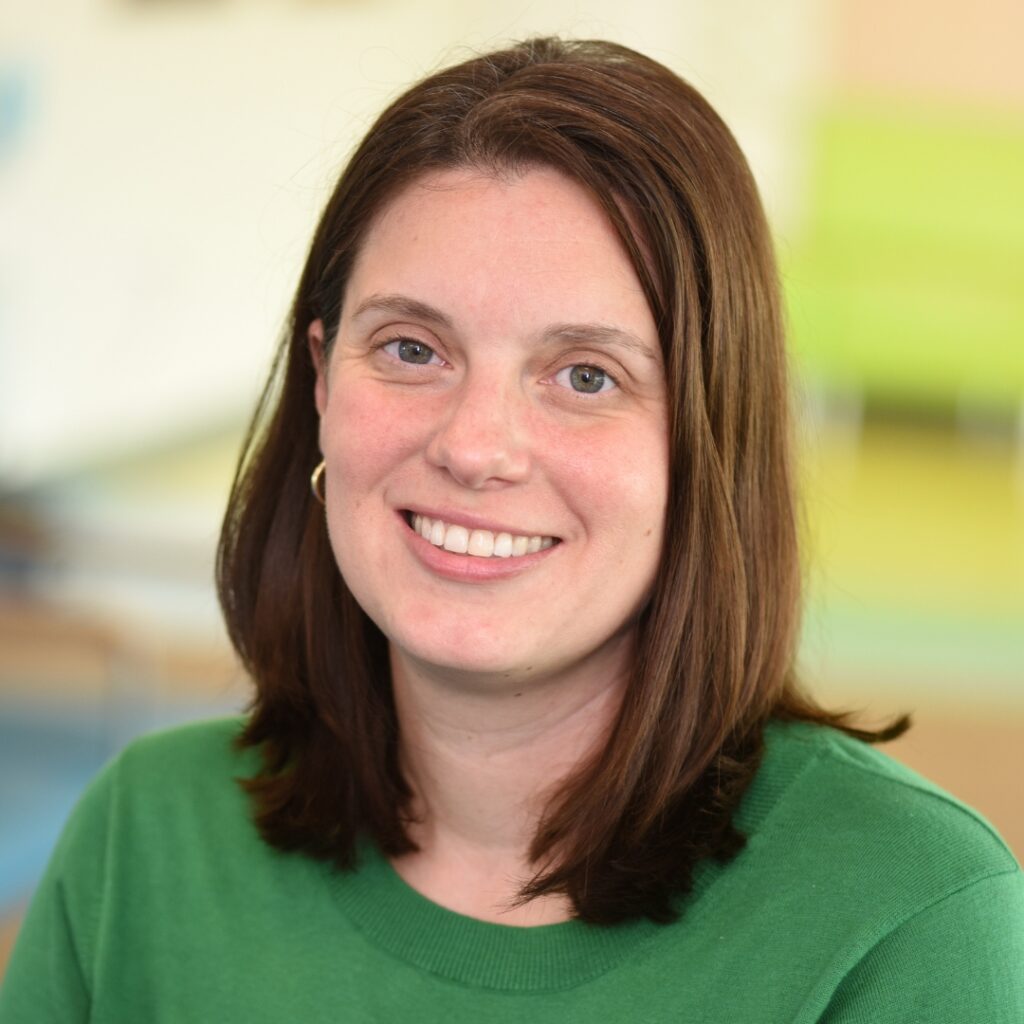 A professional headshot of a smiling woman with medium-length brown hair, wearing a green crewneck sweater and hoop earrings.
