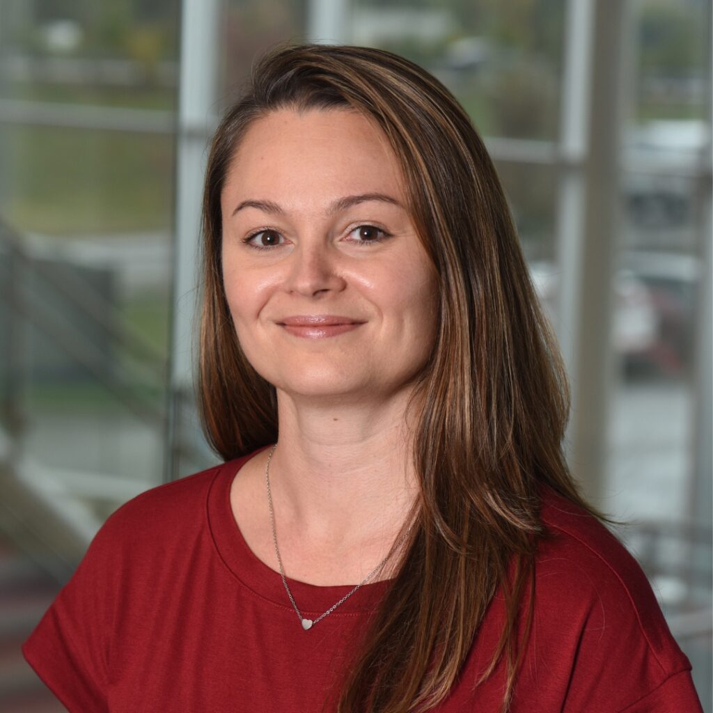 A professional headshot of a woman with long brown hair, wearing a red t-shirt and a silver heart pendant necklace smiling.
