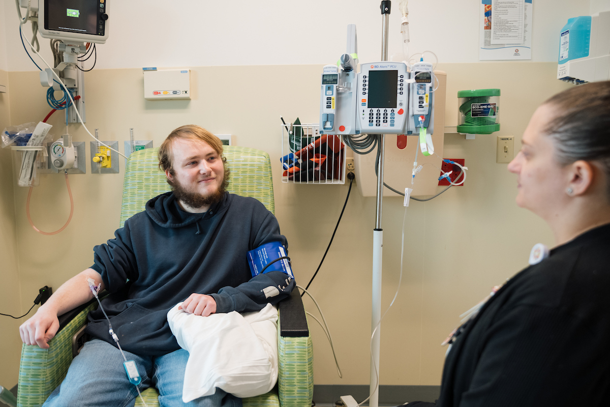 A male patient with a beard sits in a patterned chair with an IV, looking at a female healthcare worker.