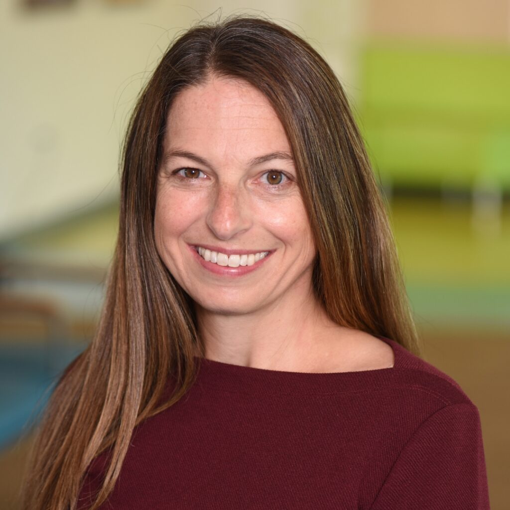 A professional headshot of a woman with long brown hair, wearing a maroon off the shoulder top.