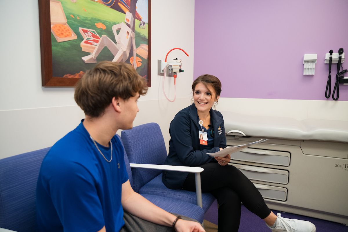 A nurse in navy scrubs smiles while talking with a teenage patient in a blue shirt inside a bright exam room with purple walls and cheerful artwork.
