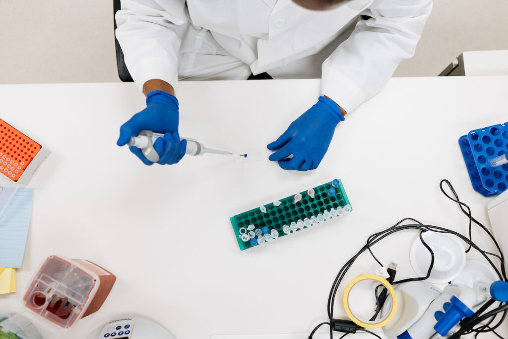 A researcher wearing blue gloves and a lab coat uses a pipette at a lab workstation with test tubes and lab equipment.
