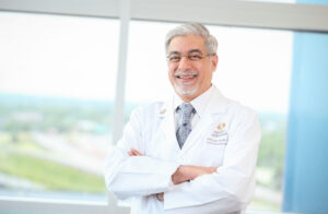 A distinguished, smiling, gray-haired male doctor in a white lab coat stands with his arms crossed in front of a window. 