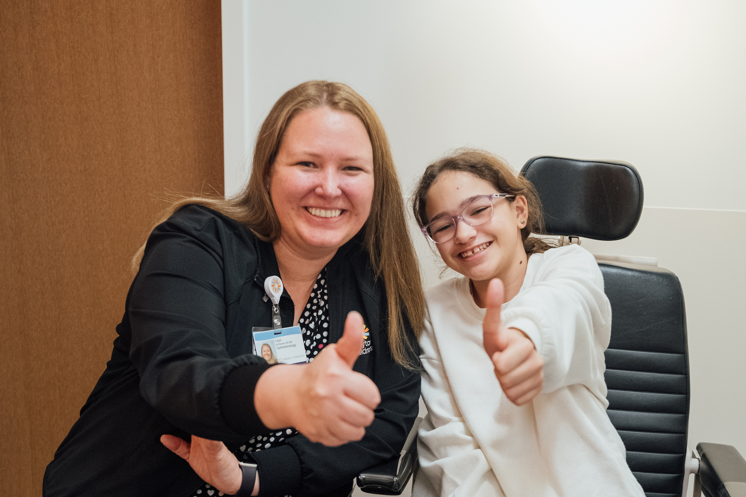 A cheerful female medical professional and a young girl in glasses both give thumbs up to the camera.