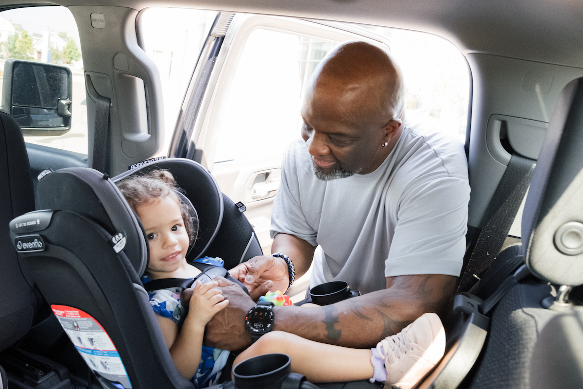 Man securing a smiling child in a car seat, adjusting straps inside a vehicle with sunlight streaming through the door.

