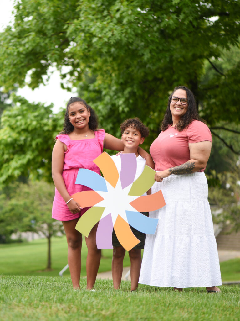 Young boy standing and smiling with his sister and mom in outdoor area.