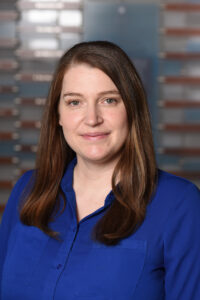 A professional headshot of a smiling woman with long brown hair, wearing a bright blue collared shirt.