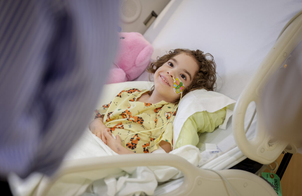young girl lays in hospital bed while smiling at visitor.
