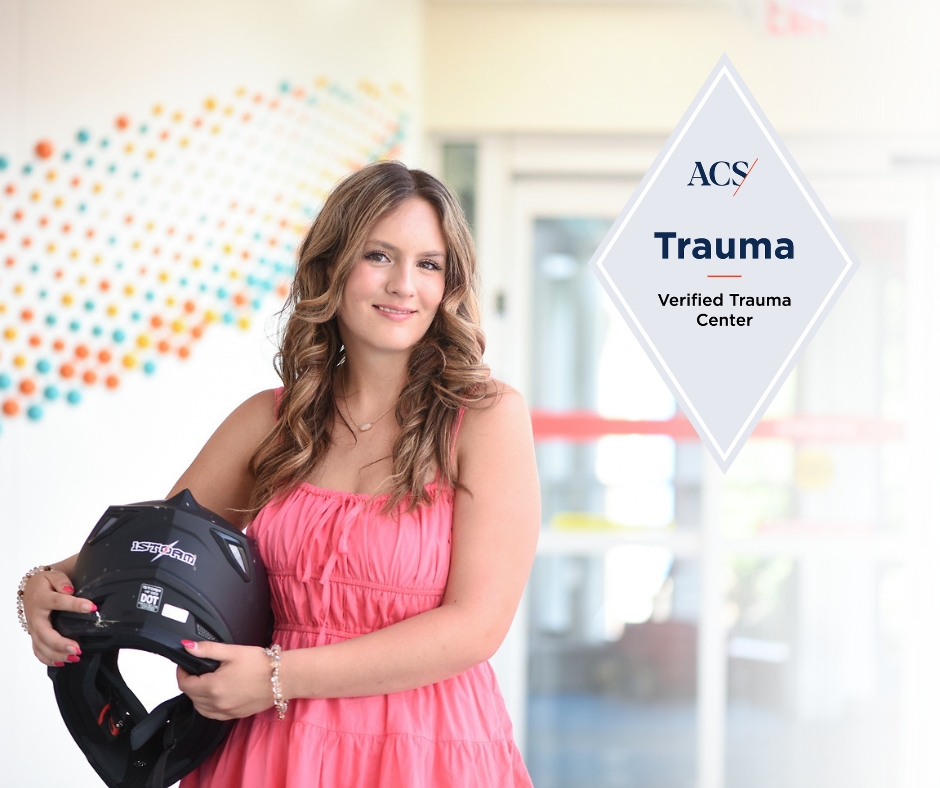  A smiling young woman in a pink dress holds a black motorcycle helmet, with a "Verified Trauma Center" logo overlay.
