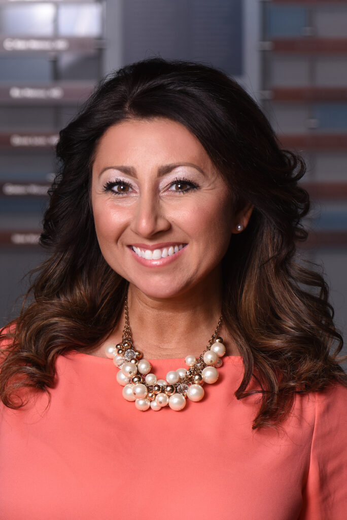 A close-up professional headshot of a smiling woman with long brown hair, wearing a coral top and a statement necklace with pearls and gold beads.