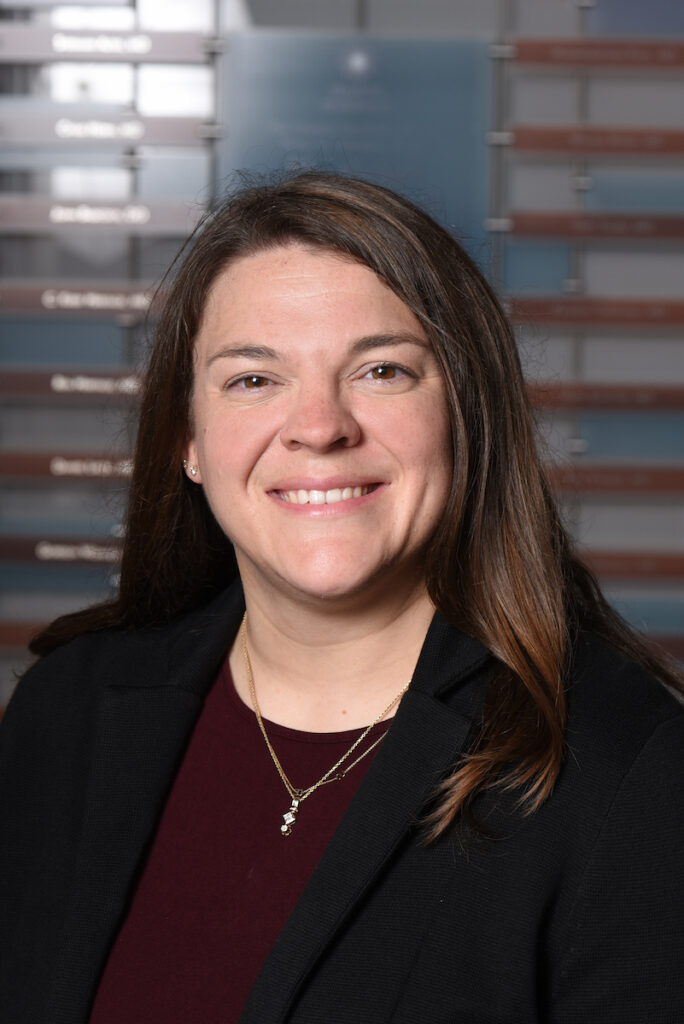 A professional headshot of a smiling woman with long brown hair, wearing a black blazer over a burgundy top and a delicate necklace.