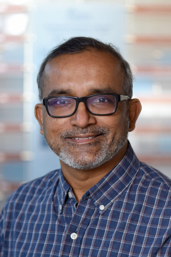 A professional headshot of a smiling man with a gray beard and glasses, wearing a blue plaid shirt.