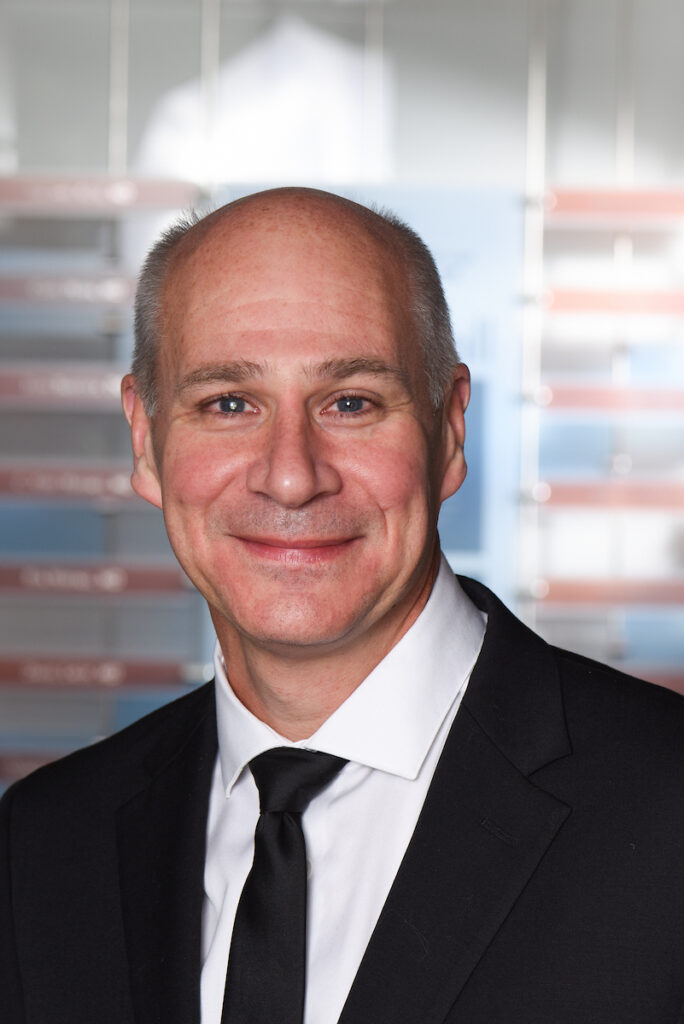  A professional headshot of a smiling man in a black suit and tie, looking directly at the camera.