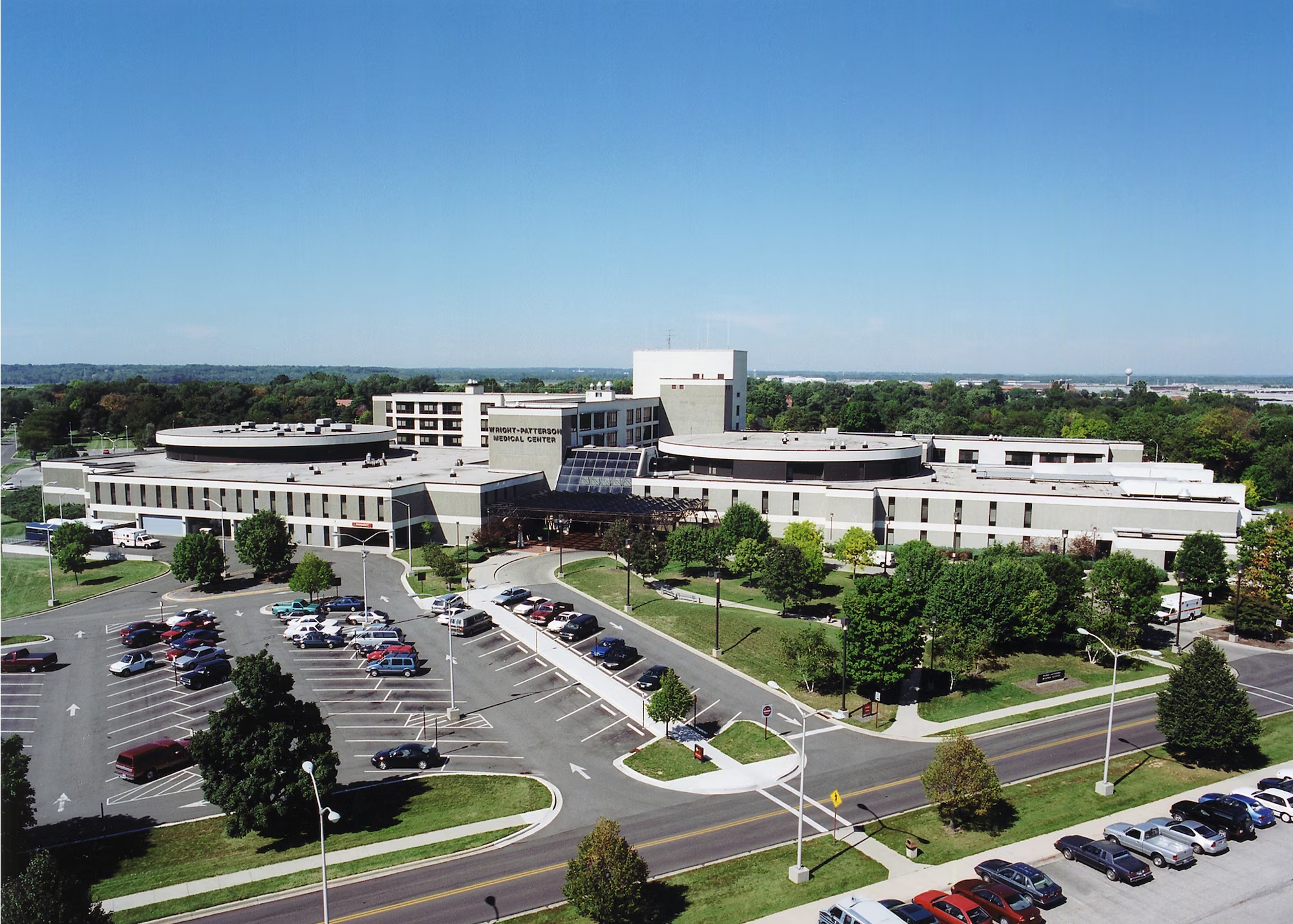 An aerial view of the sprawling Wright-Patterson Medical Center, a multi-building complex with large parking lots, surrounded by trees under a clear blue sky.