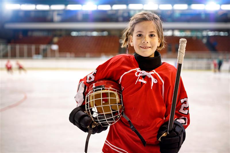 Young ice hockey player holds hockey gear while on the ice rink.