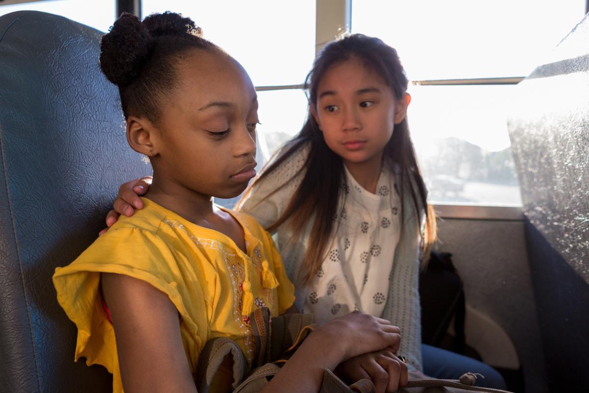 two young girls sit on a bus, one helps comfort the other who has experienced bullying