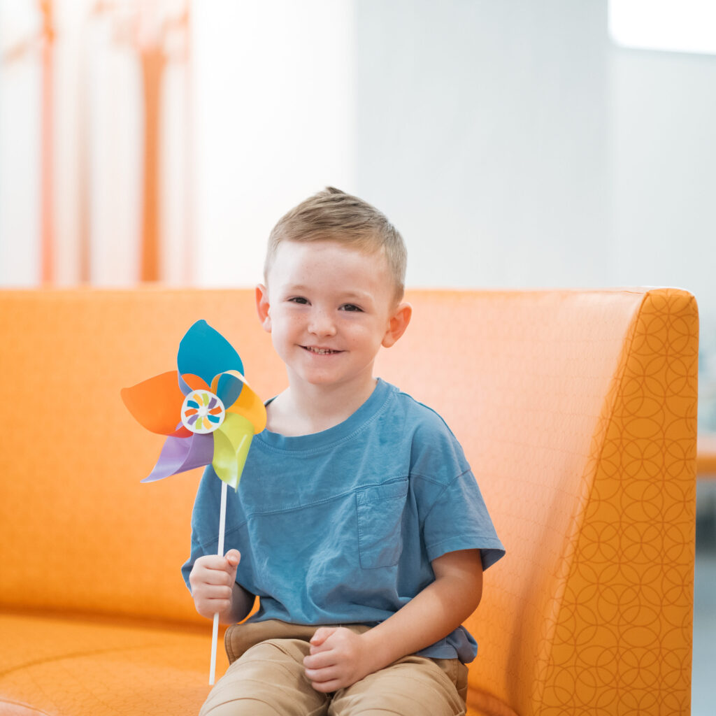 Young child sitting on orange couch in bright space while smiling and holding a colorful pinwheel toy.