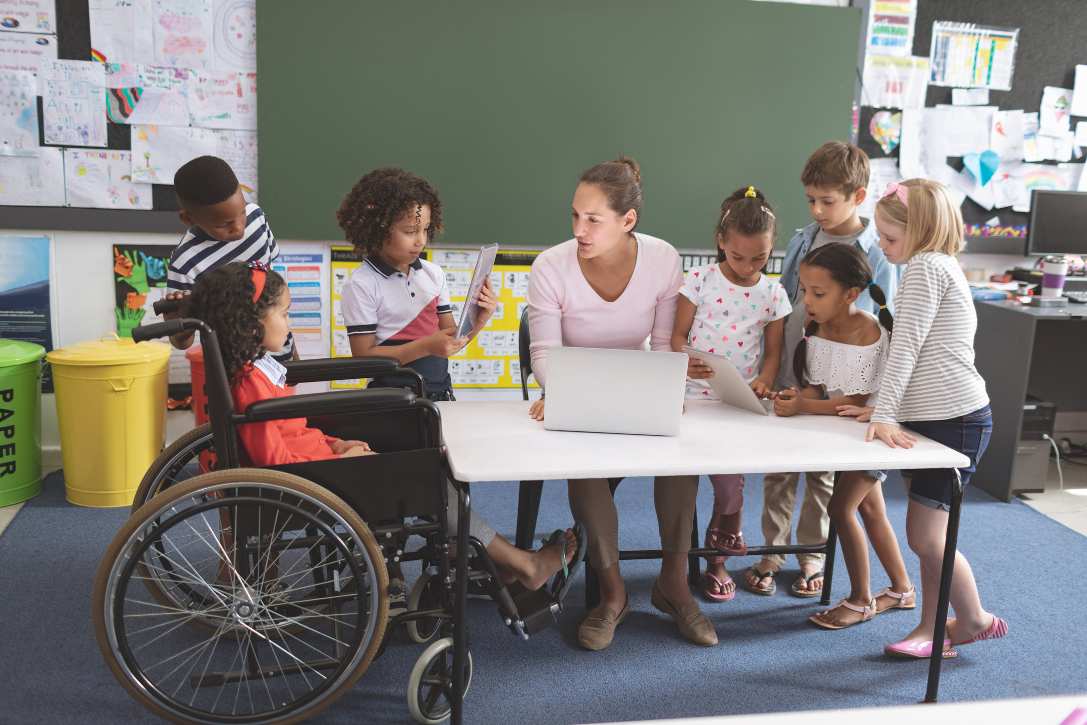 a teacher sits at a desk surrounding by several students in school