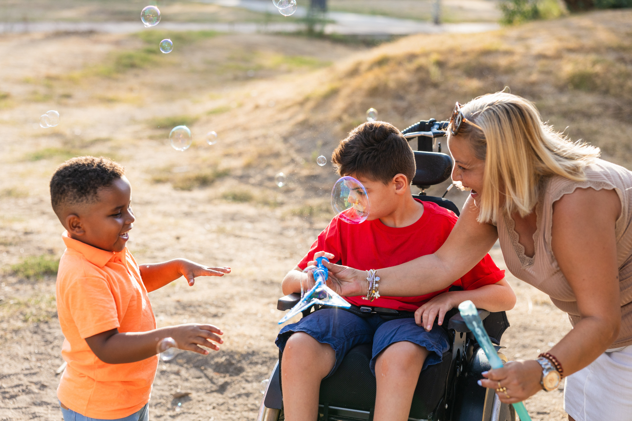 young boy in wheelchair plays with bubbles alongside his friend outside