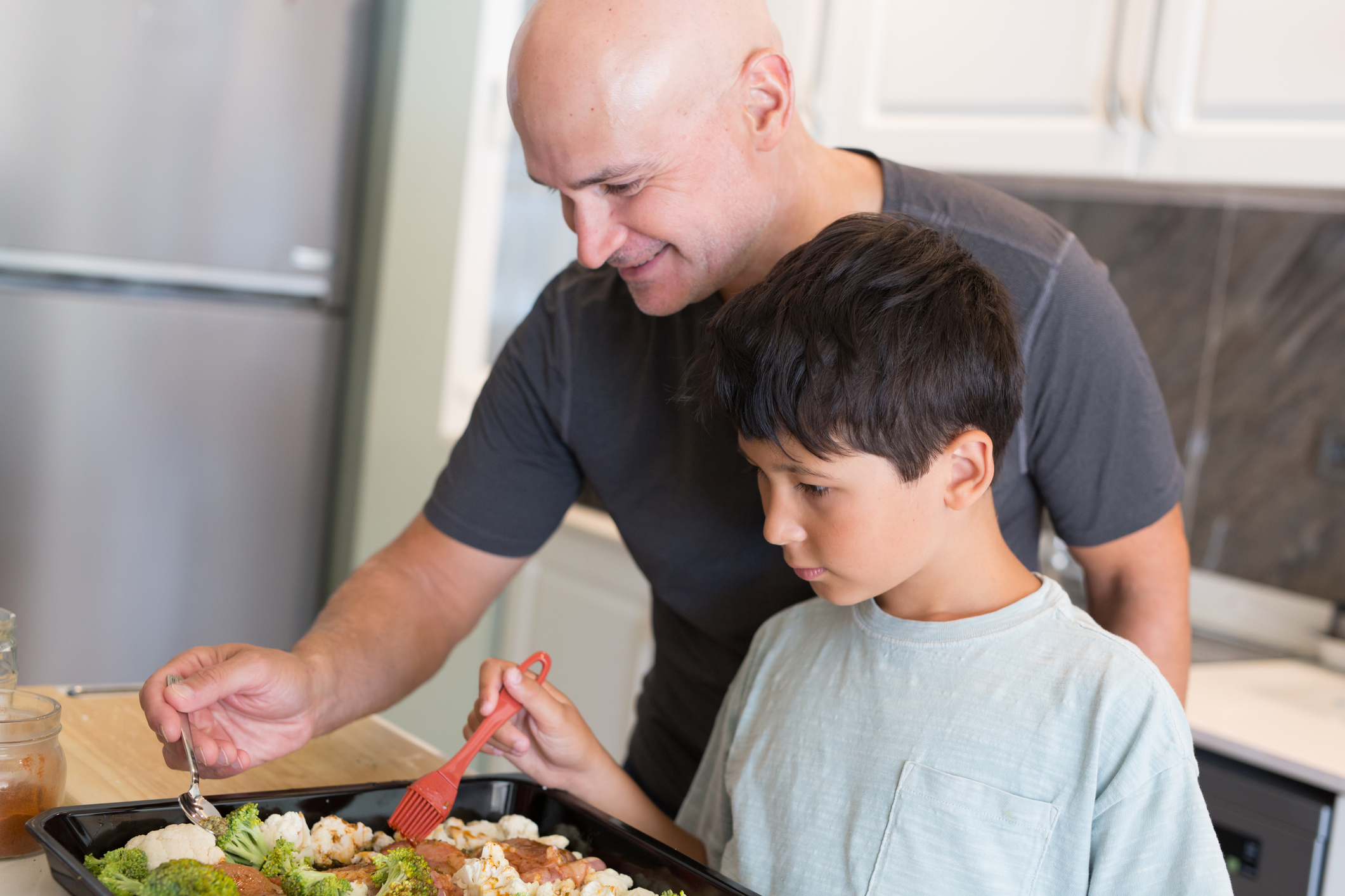 Father and son put garlic and herbs on a chicken and vegetable dish they are preparing to cook