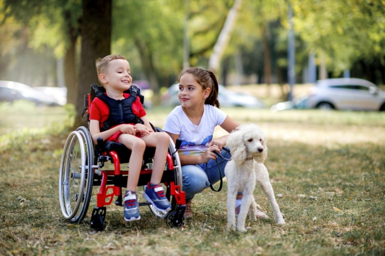 Boy in wheelchair smiles at a park with his friend and a dog