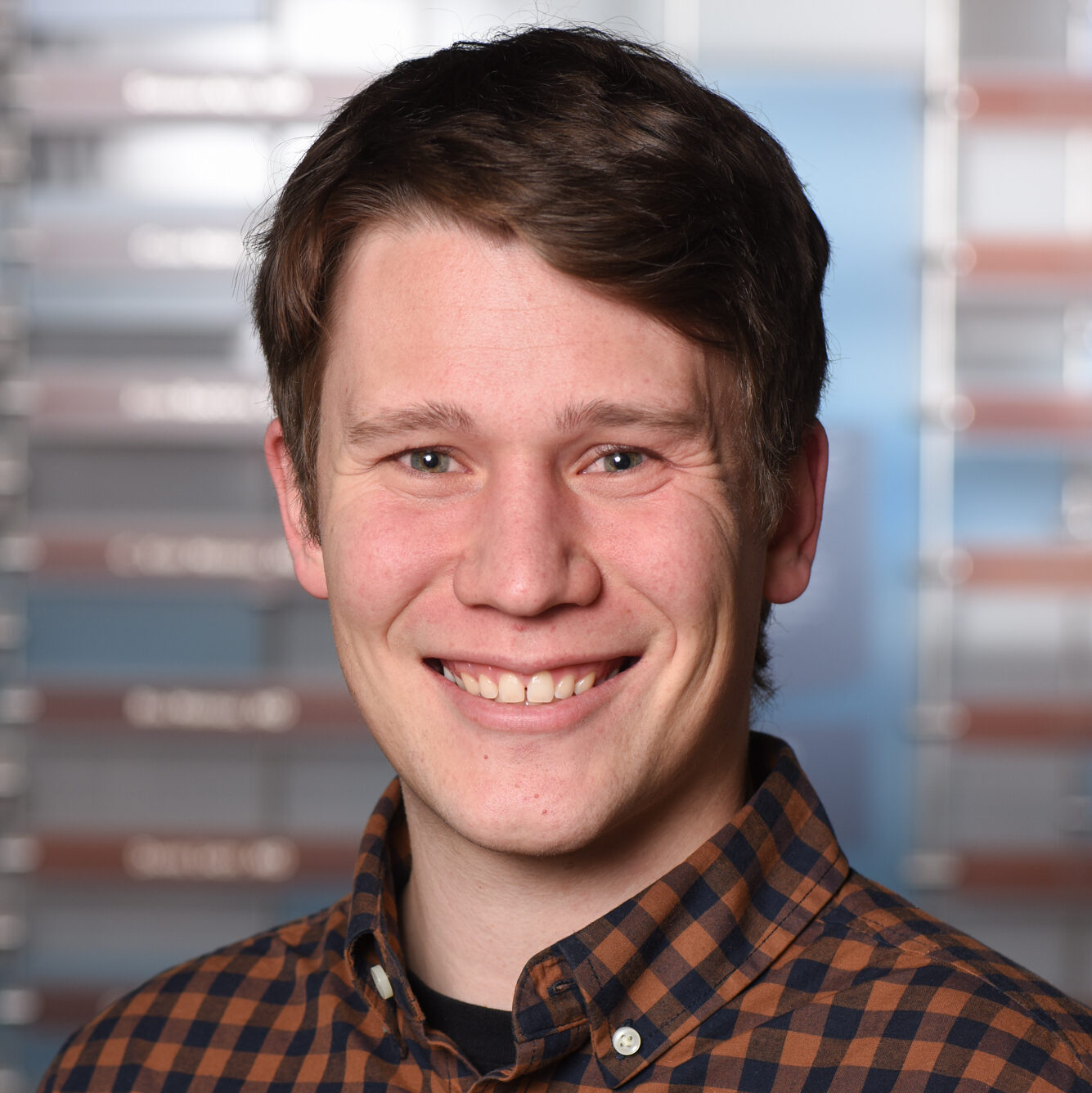 A professional headshot of a smiling man with short brown hair, wearing a orange and black plaid collared shirt.