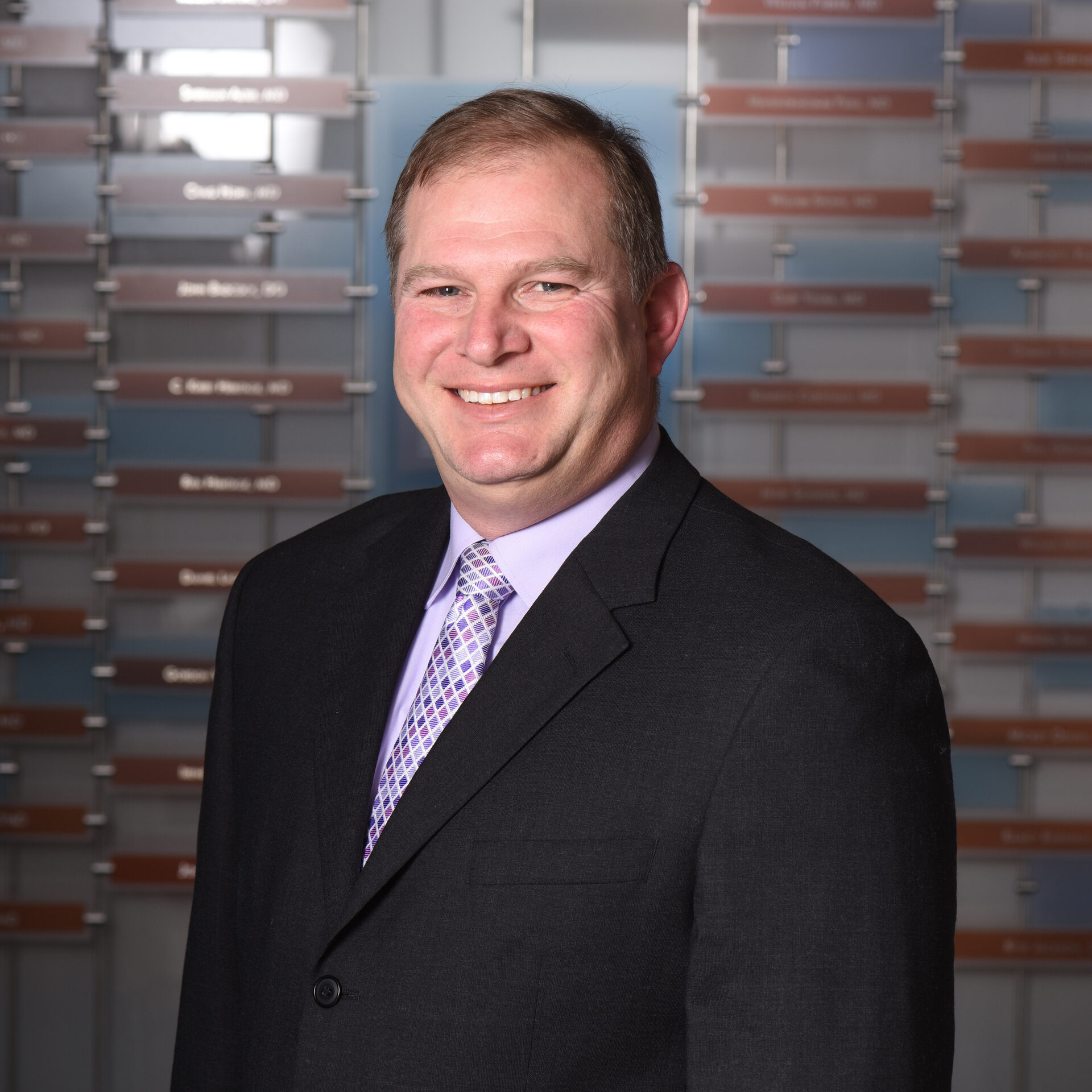 A professional headshot of a smiling man with short hair, wearing a black suit jacket with a purple collared shirt and purple tie.