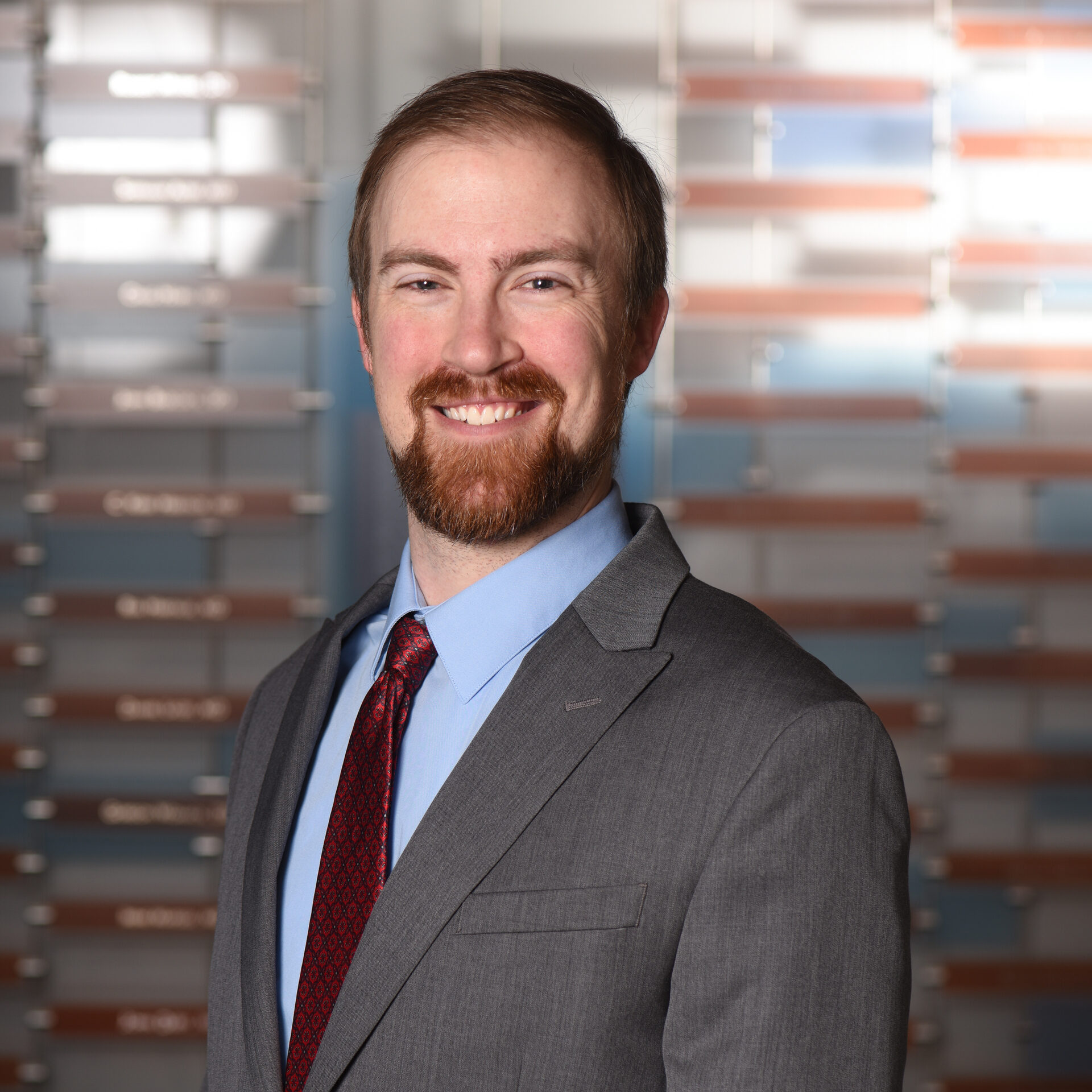 A professional headshot of a smiling man with short hair, wearing a grey suit jacket with a blue collared shirt and red tie.