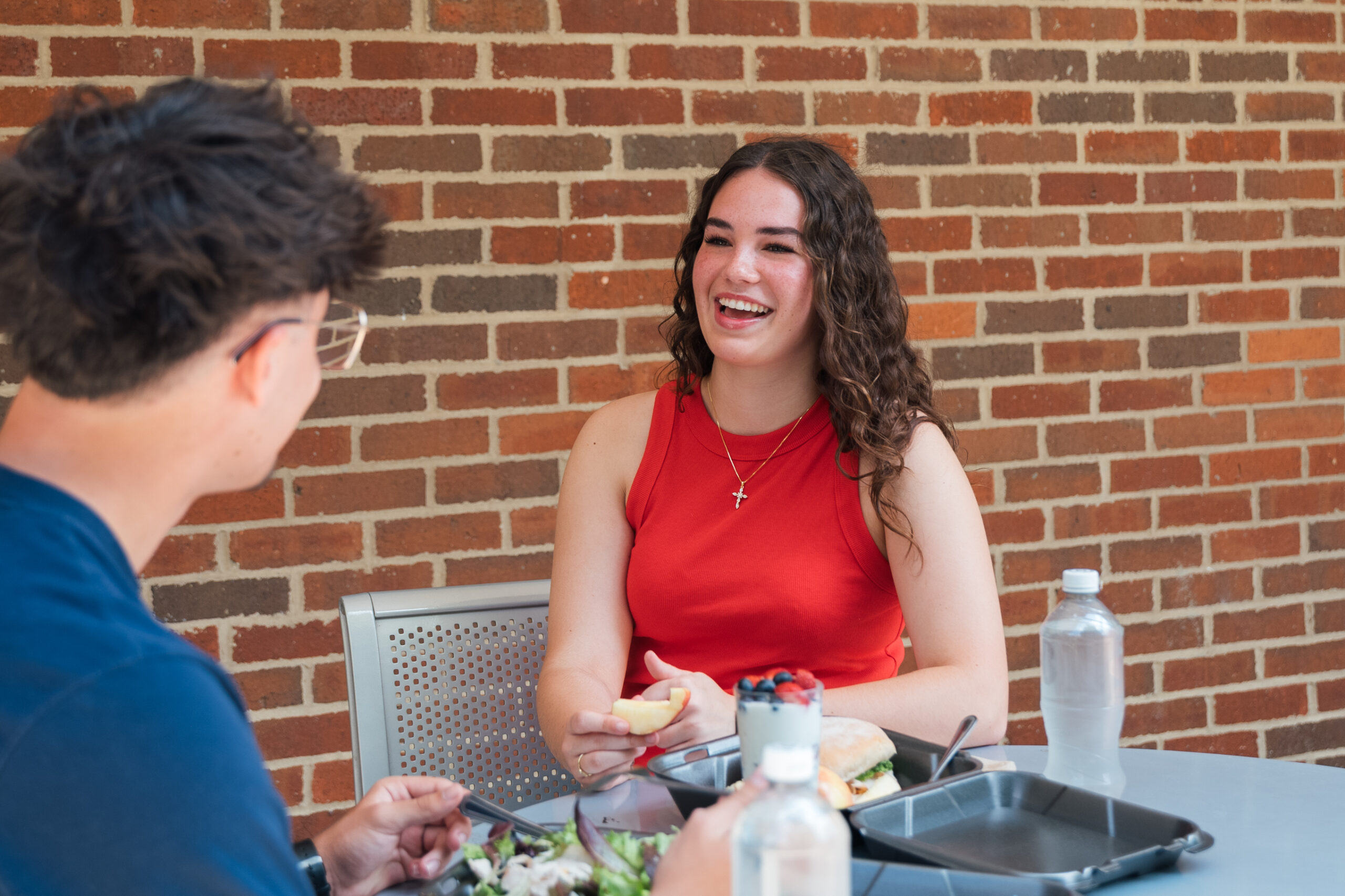 Teen girl with long curly dark hair talks and eats lunch outside with a friend at college.