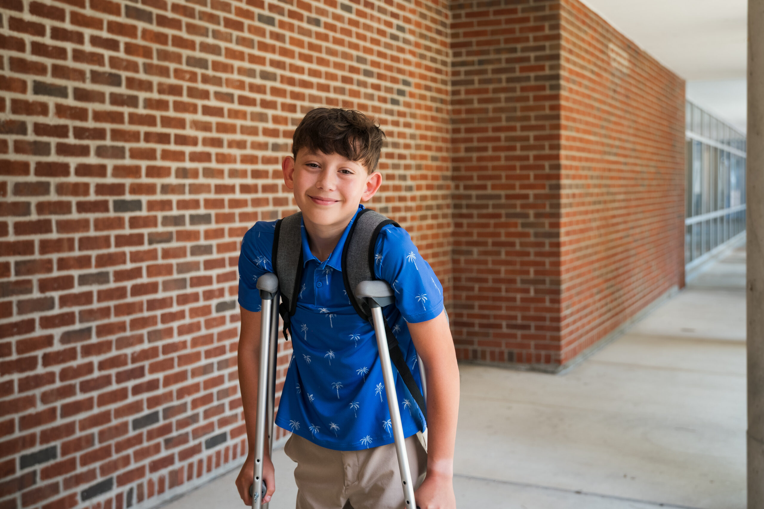 Middle school boy wearing a blue shirt walks into school with a backpack while using crutches.
