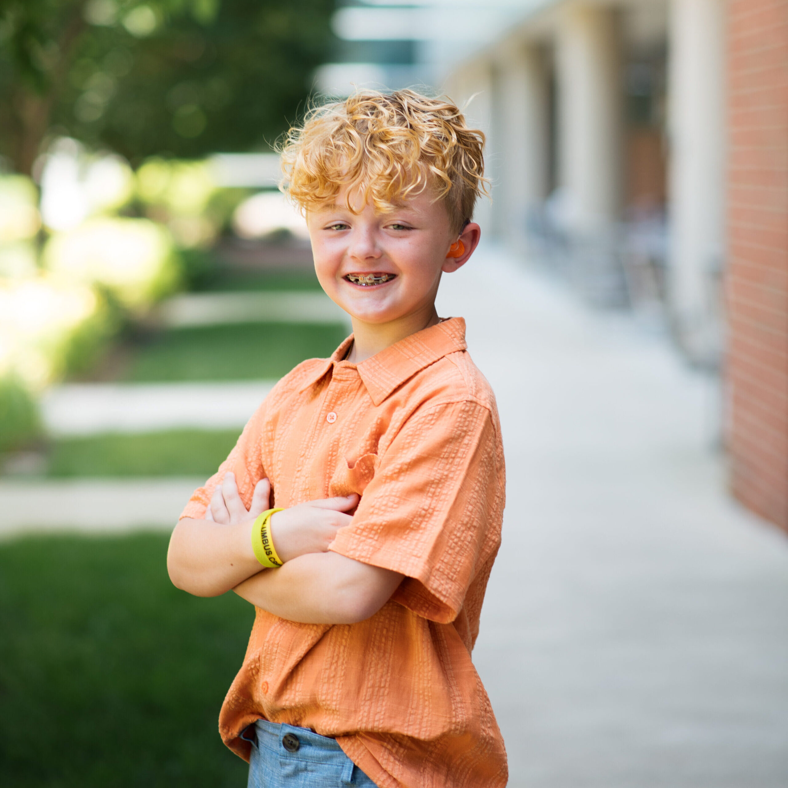 Young boy with blonde curly hair in an orange shirt with arms crossed smiles while standing outside of Dayton Children's.