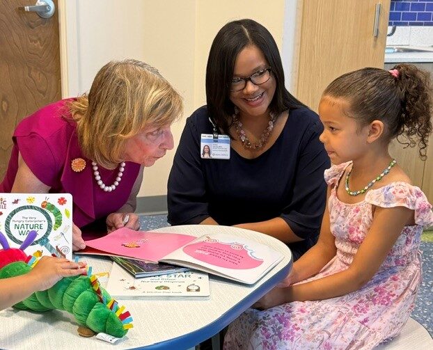 First Lady Fran DeWine sits at a small table reading a children's book provided by the Imagination Library of Montgomery County Fund with a young girl in a pink dress with her mom smiling next to her.