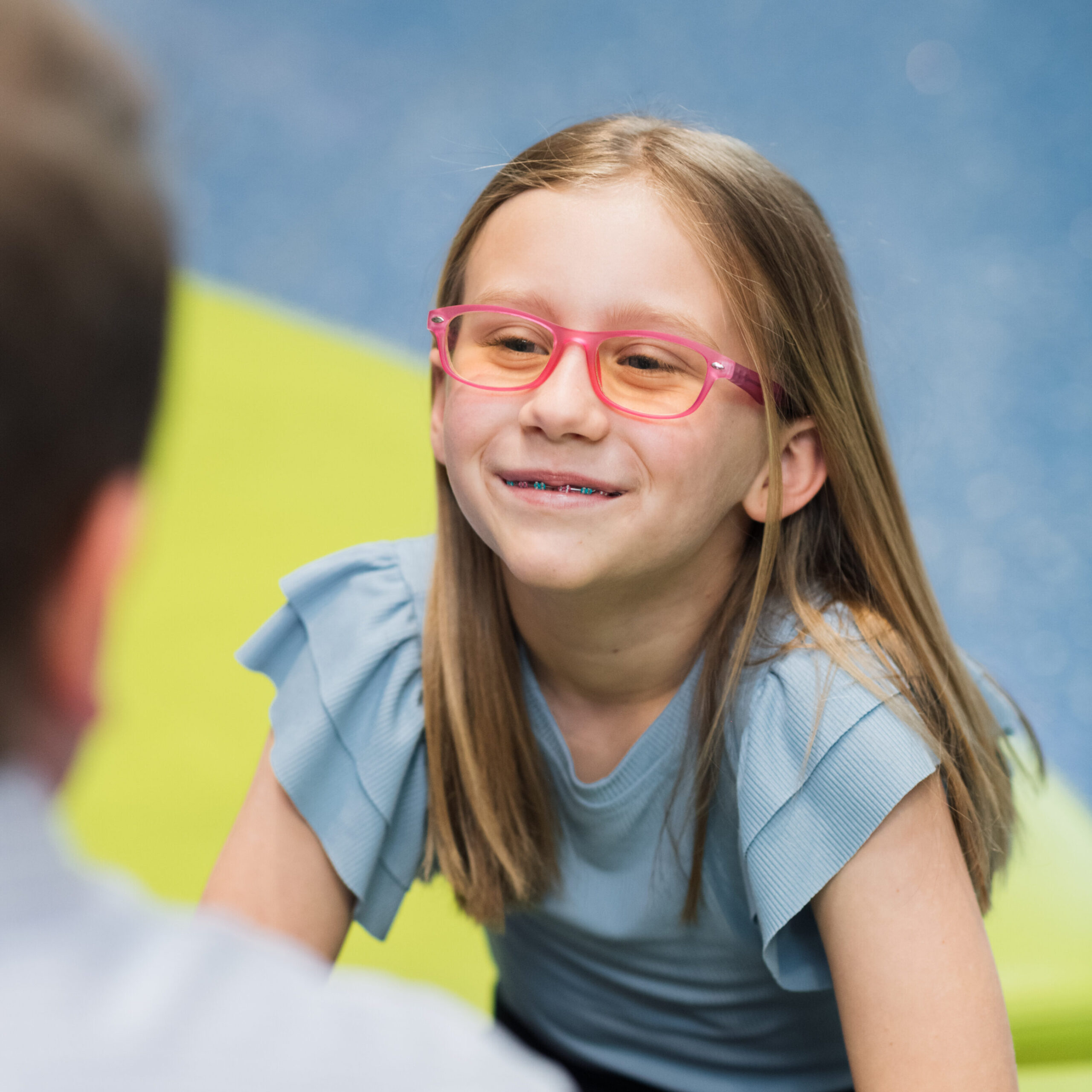A girl wearing glasses, sits smiling on a bench.