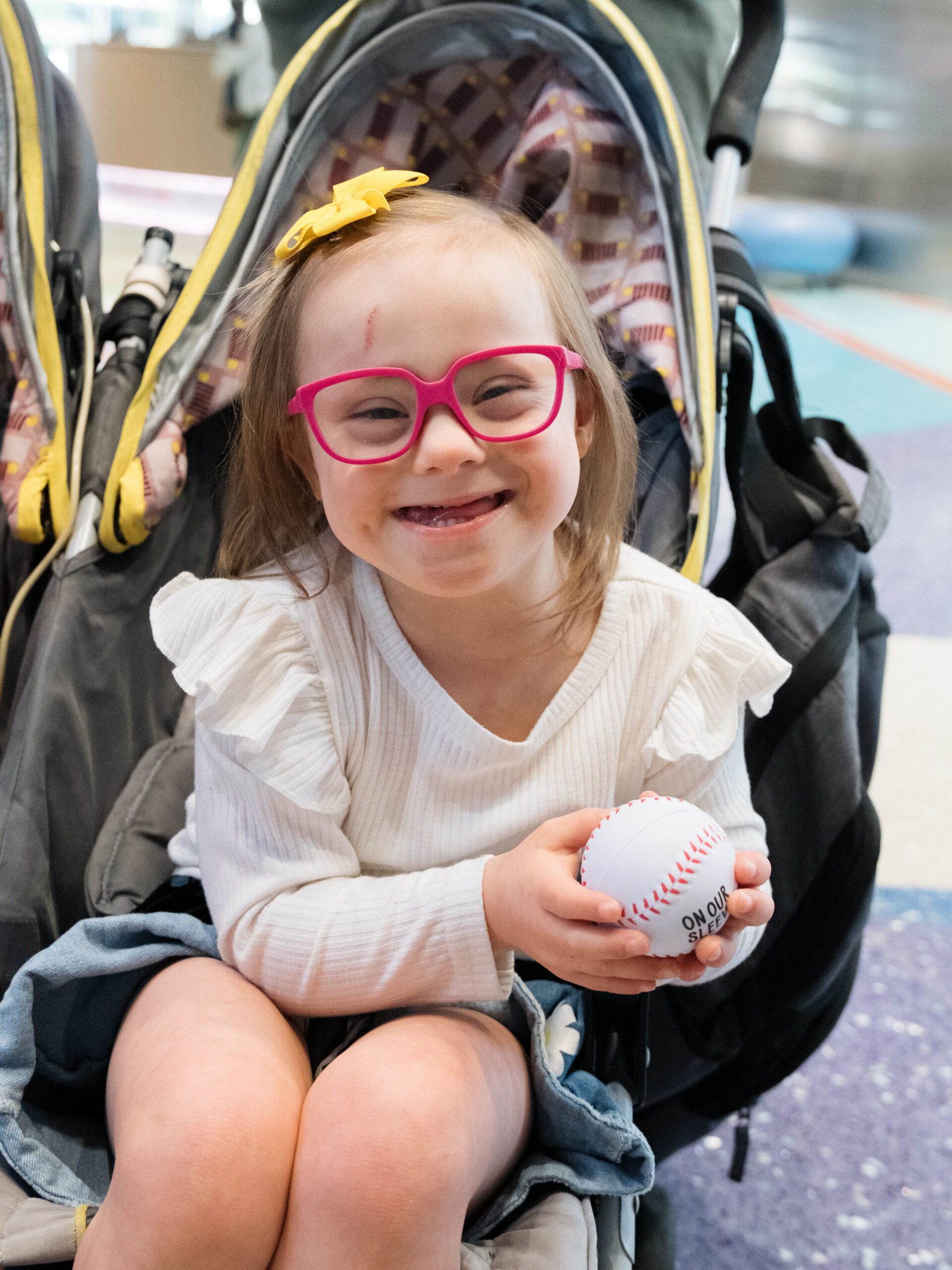 A young girl with Down Syndrome smiles at the camera while holding a foam baseball.