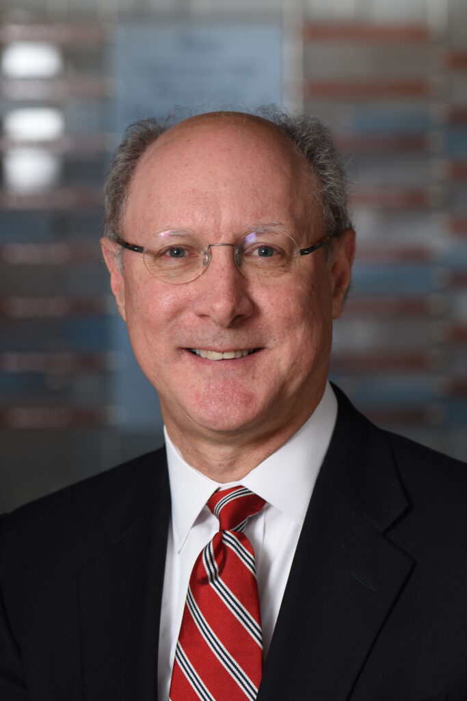 Headshot of Charles Kidwell smiling and wearing glasses and a red tie with a dark suit jacket.