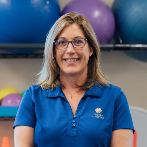 A friendly woman wearing glasses and a blue Dayton Children's polo shirt smiles in a gym setting with large exercise balls behind her.