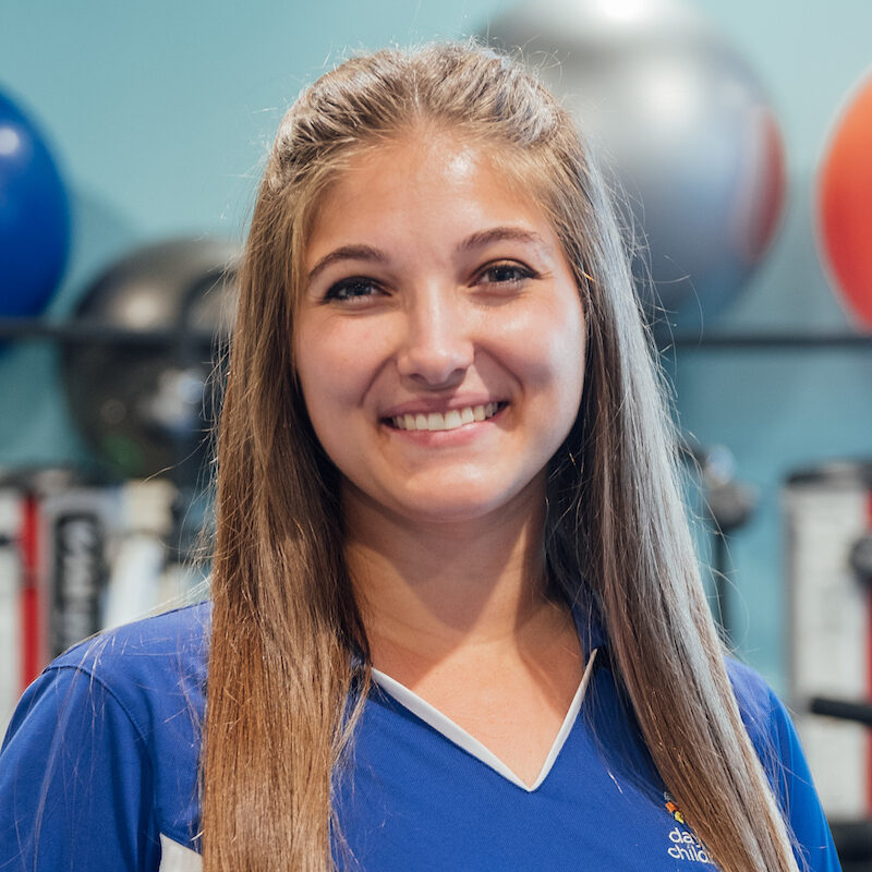 A smiling young woman with long, straight brown hair, wearing a blue and white athletic polo shirt, in a therapy gym setting.