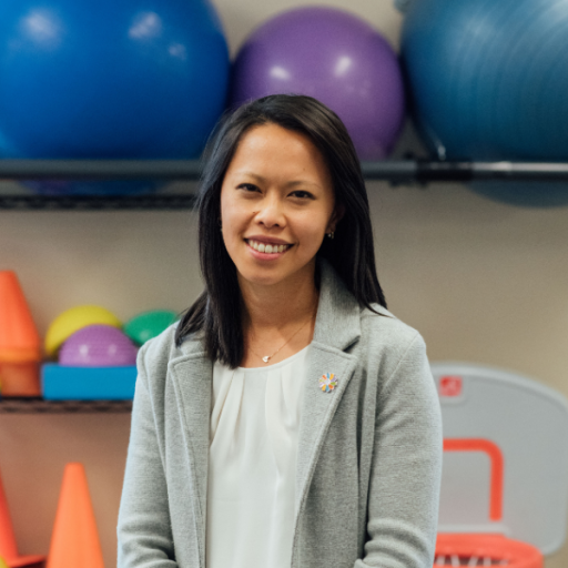 A woman smiles in a gray blazer over a white blouse, wearing a small Dayton Children's pin, in a therapy gym setting.